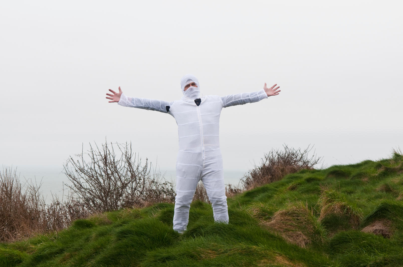 Man in a tight white boiler suit with his head in white bandages standing in a field with his arms outstretched