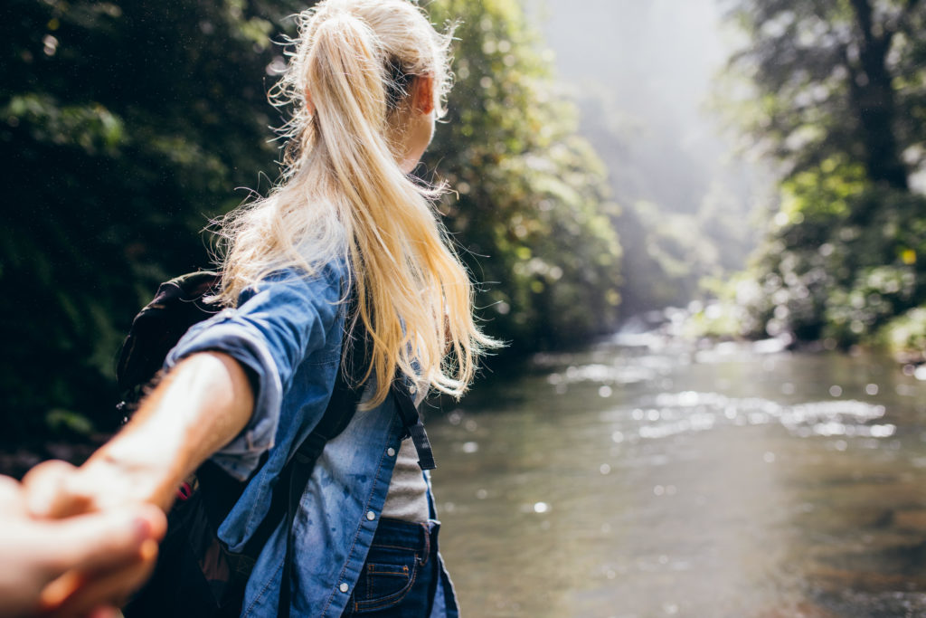 Young woman leading her boyfriend on the forest hike. Point of view shot of couple crossing the creek holding hands.