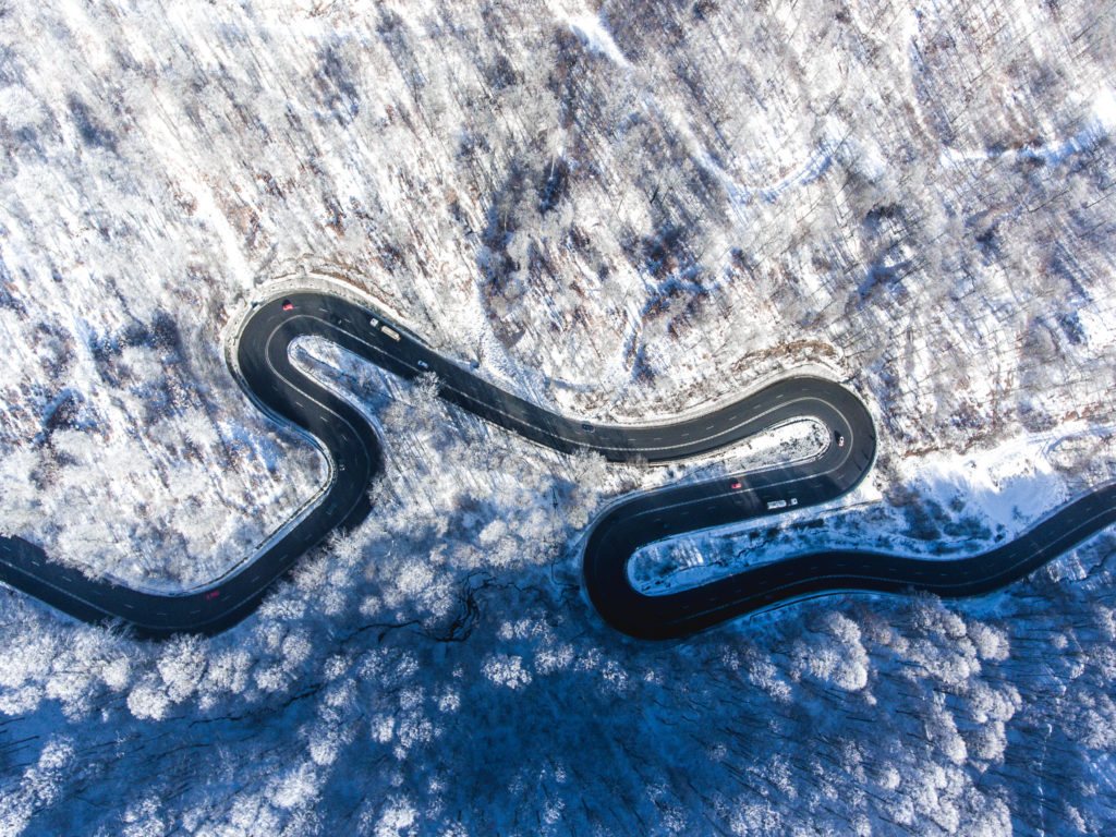 Aerial drone view of a curved winding road through the forest high up in the mountains in the winter with snow covered trees